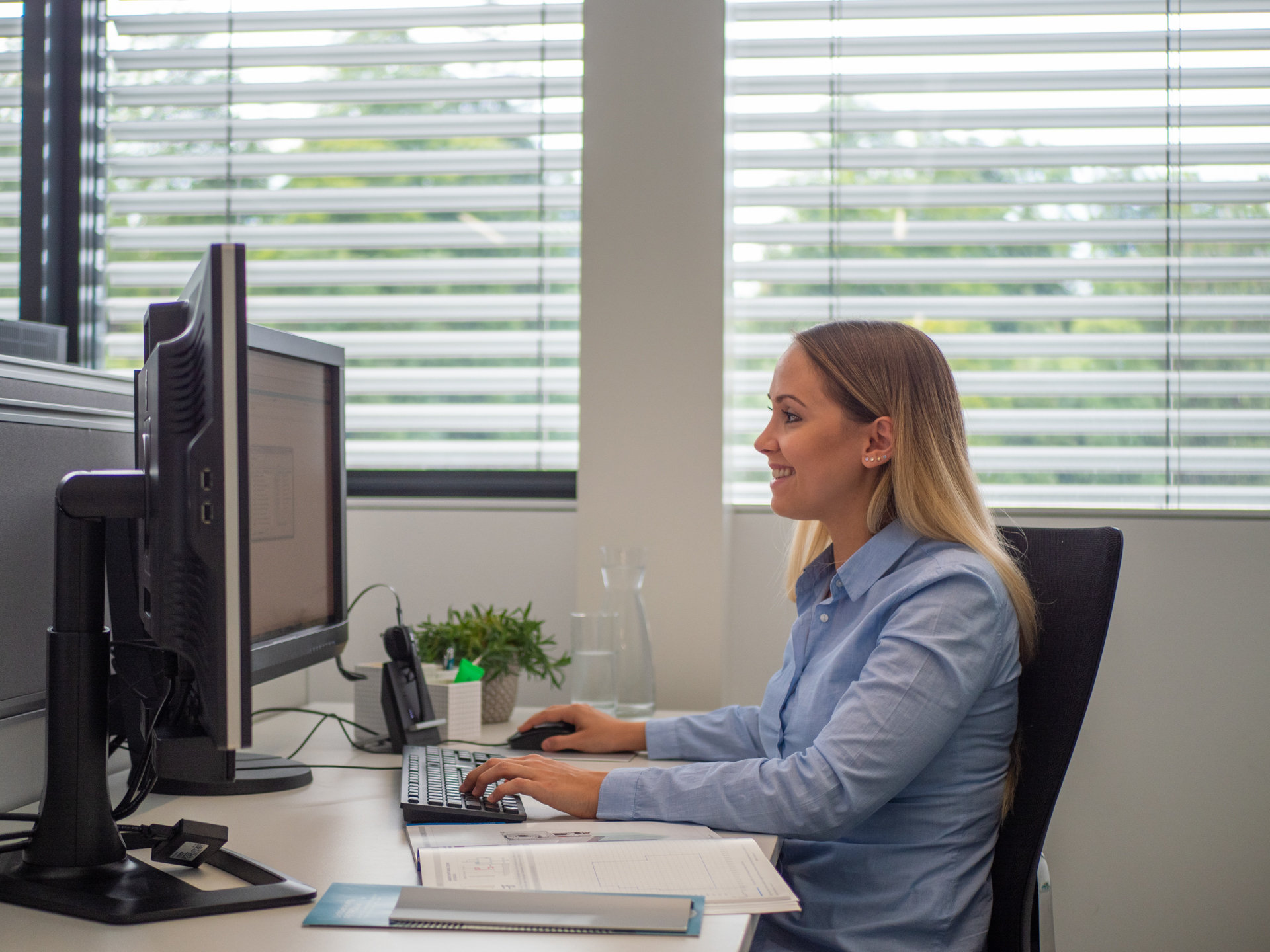 Frau sitzt am Schreibtisch und arbeitet am PC, im Hintergrund ist Sonnenschutz zu sehen.